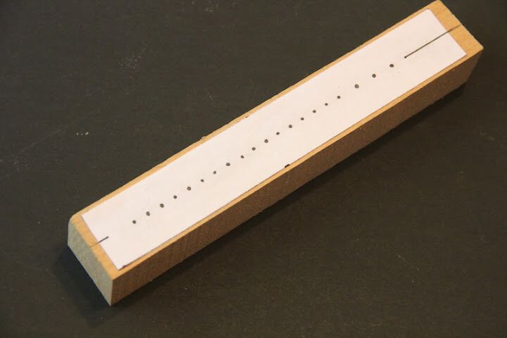 Figure 2 — Drilling a John Smith tracker bar on a drill press against a fence, each hole set from a cumulative-dimension datum with a steel rule and a brad-point bit, backed to prevent breakout.